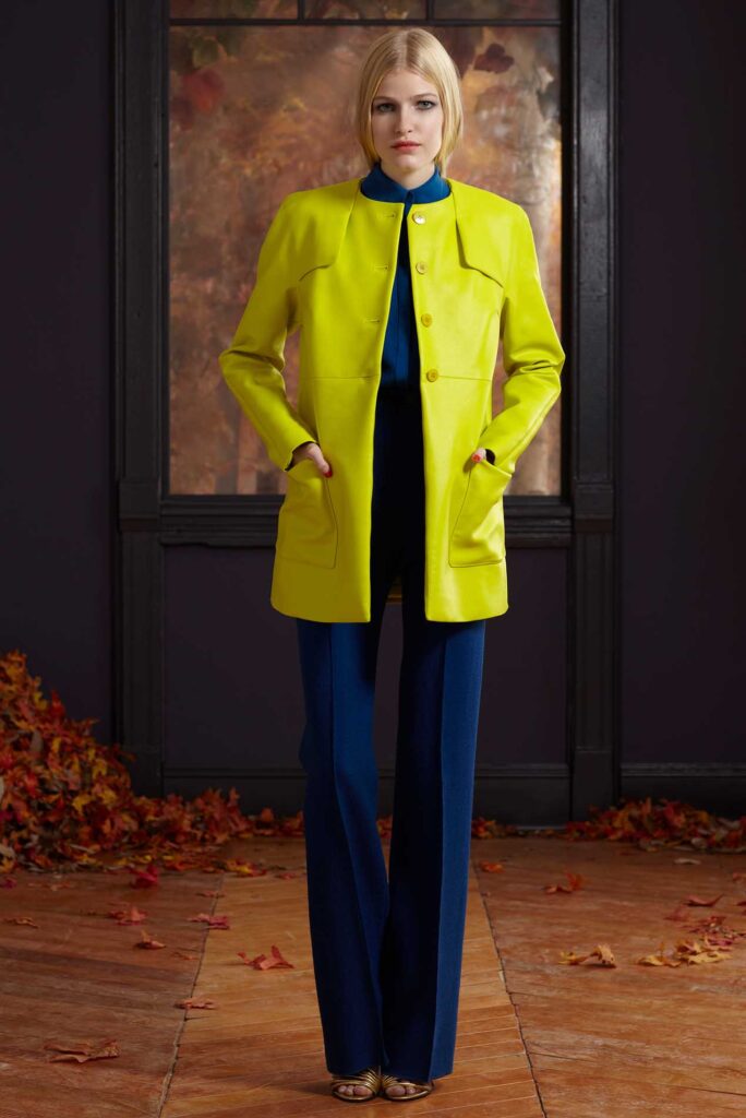 Woman in yellow coat over blue outfit stands indoors on leaf-strewn floor, autumn scene outside window.