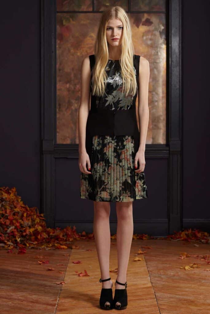 Woman in a black floral dress and black heels stands indoors with autumn leaves scattered on the floor.