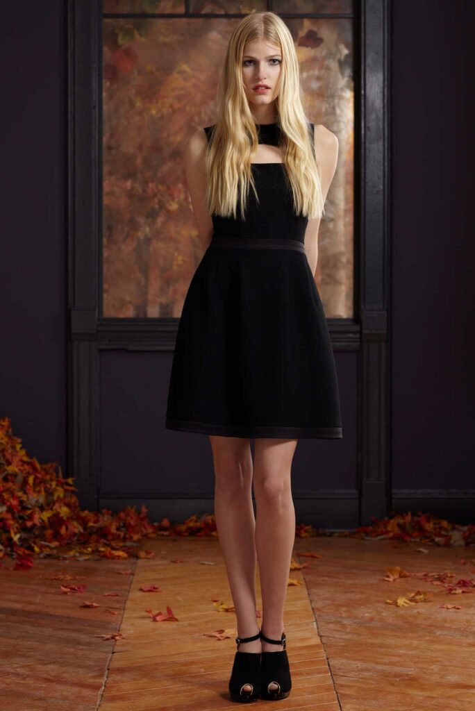 Woman in a sleeveless black dress stands indoors, autumn leaves scattered on the floor behind her.
