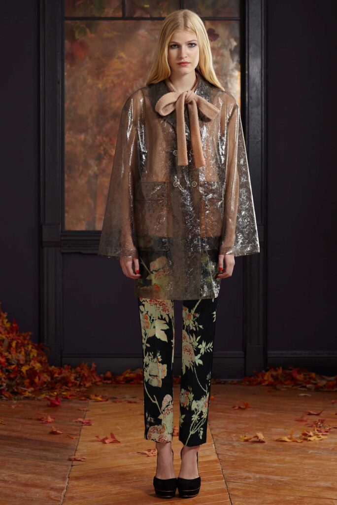 A woman wears a sheer coat, tan bow blouse, floral pants, black heels, standing on a leaf-strewn floor.