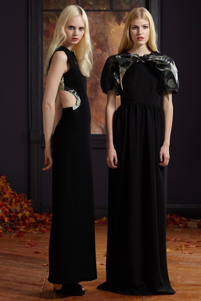 Two women in long black dresses stand indoors, with autumn leaves on the floor and a window behind them.