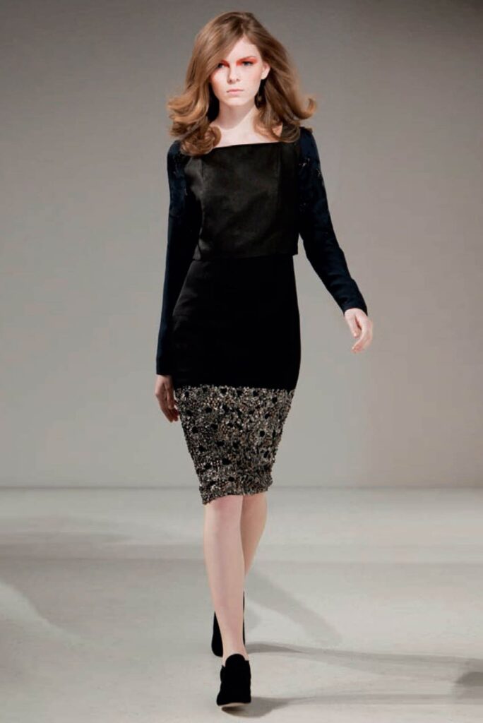 Model walks runway in a black long-sleeve dress with a sparkly silver skirt and black heels.