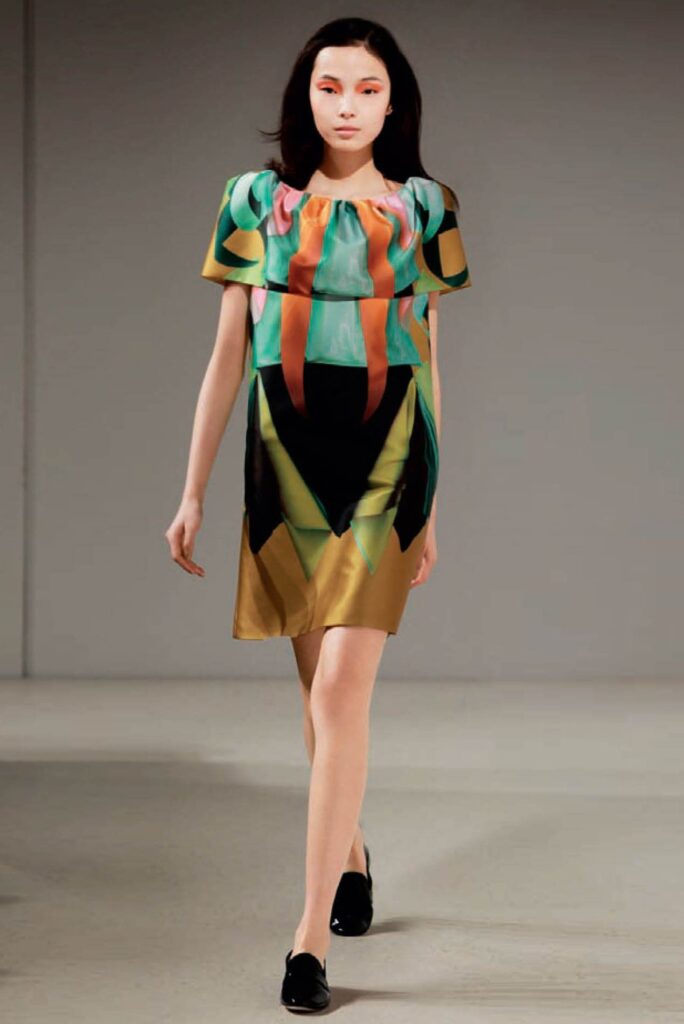 Model wearing a colorful geometric dress and black shoes walking on a runway in a minimalist room.