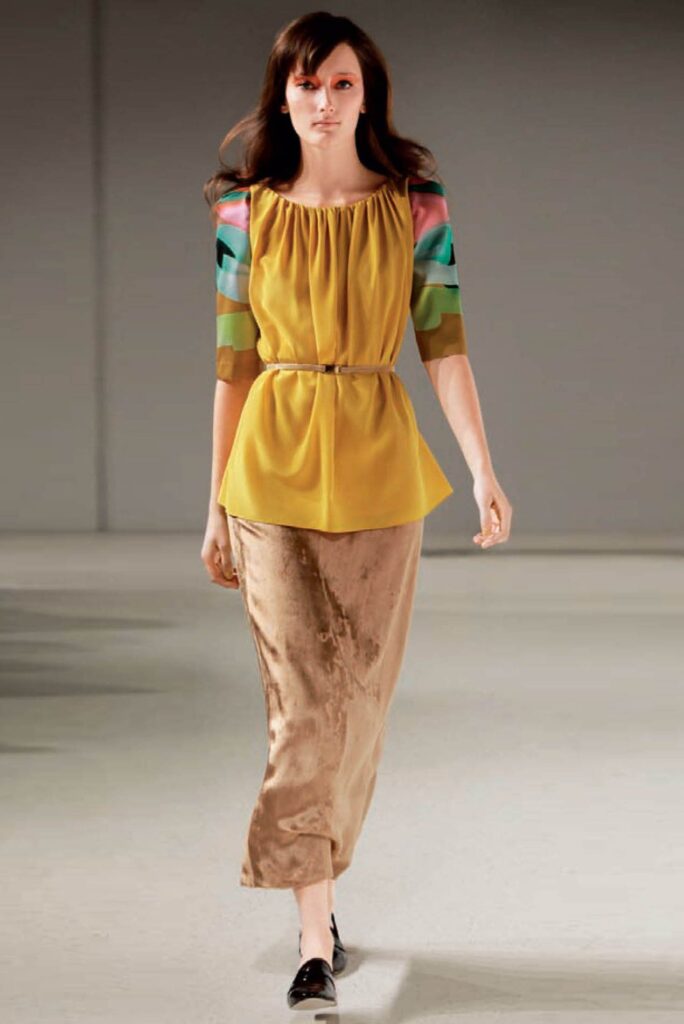 Model in a yellow belted top with colorful sleeves, beige skirt, and black shoes on a runway.