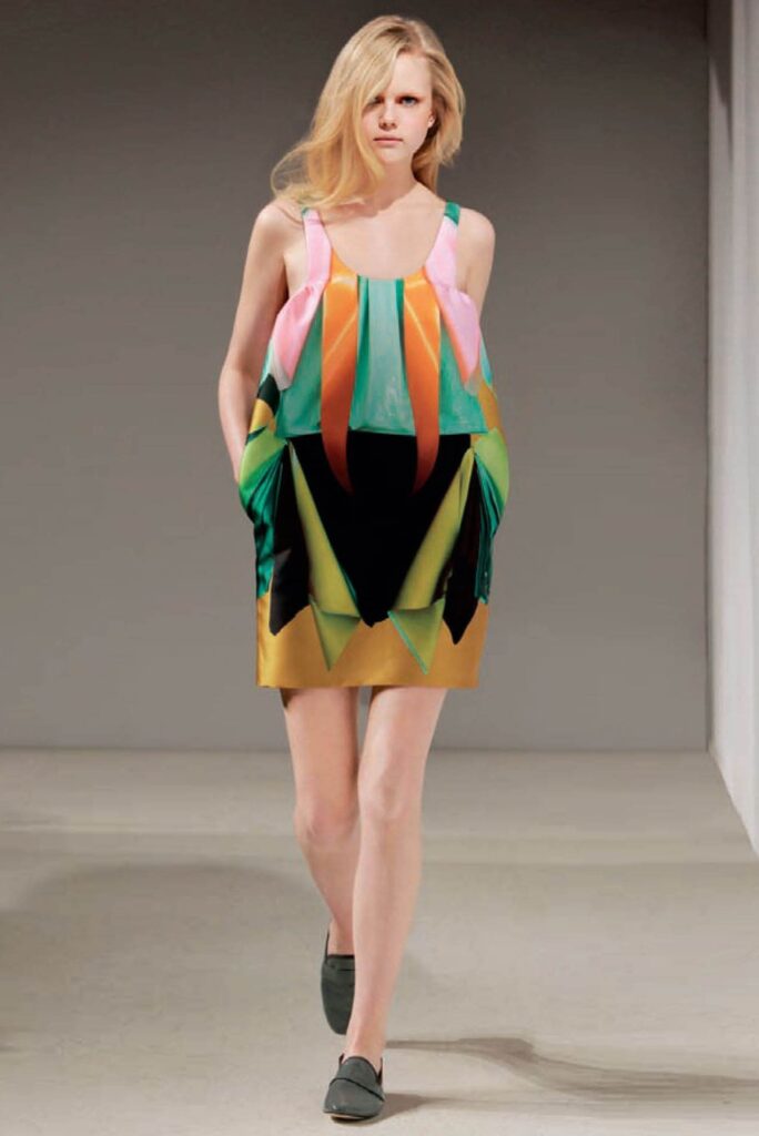 Model in a colorful geometric dress with pockets, walking on a runway with neutral background.
