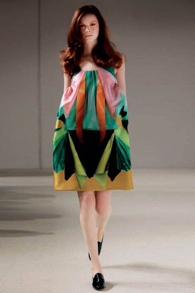 Model in a loose, colorful geometric dress with black shoes, walking on a plain indoor runway.