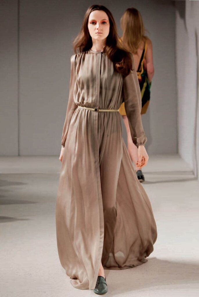 Model in a flowing taupe gown with long sleeves walks on a fashion runway; another model is behind her.