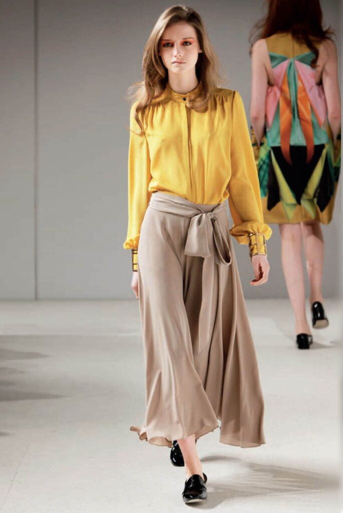 Woman on runway wears yellow blouse, beige wide-leg pants, and black shoes; another model in colorful dress.