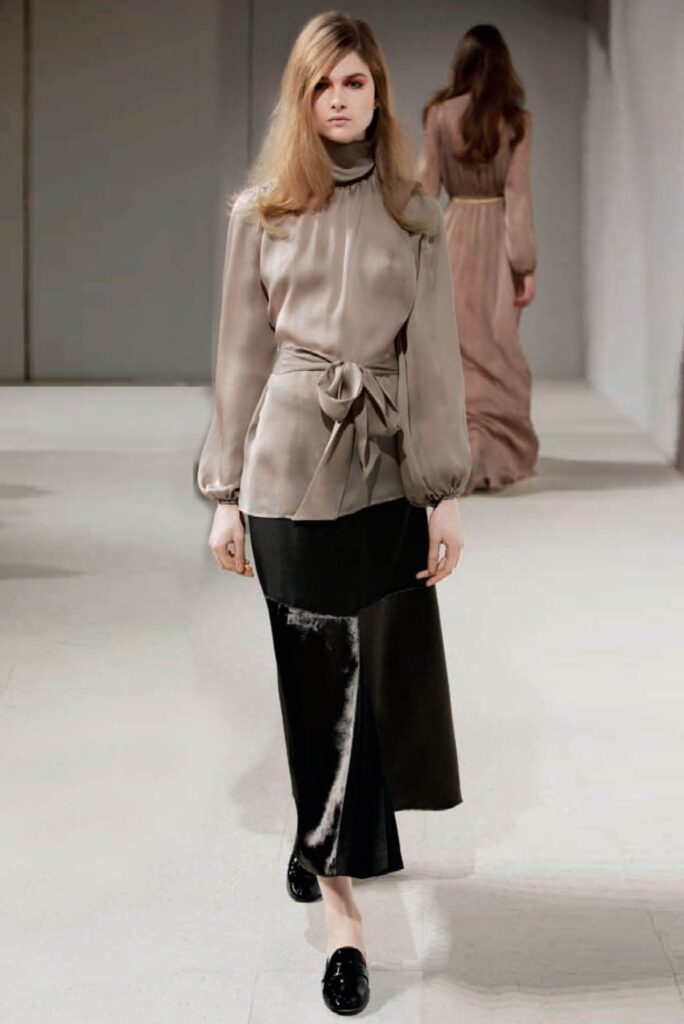 Model in taupe blouse and black skirt walks runway; another model in a beige dress walks ahead.