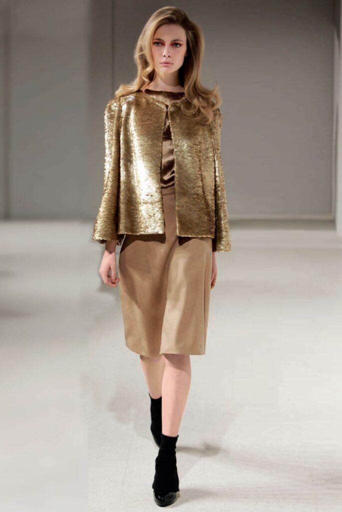Model wears a gold sequin jacket over a beige dress with black socks and heels, walking a runway.