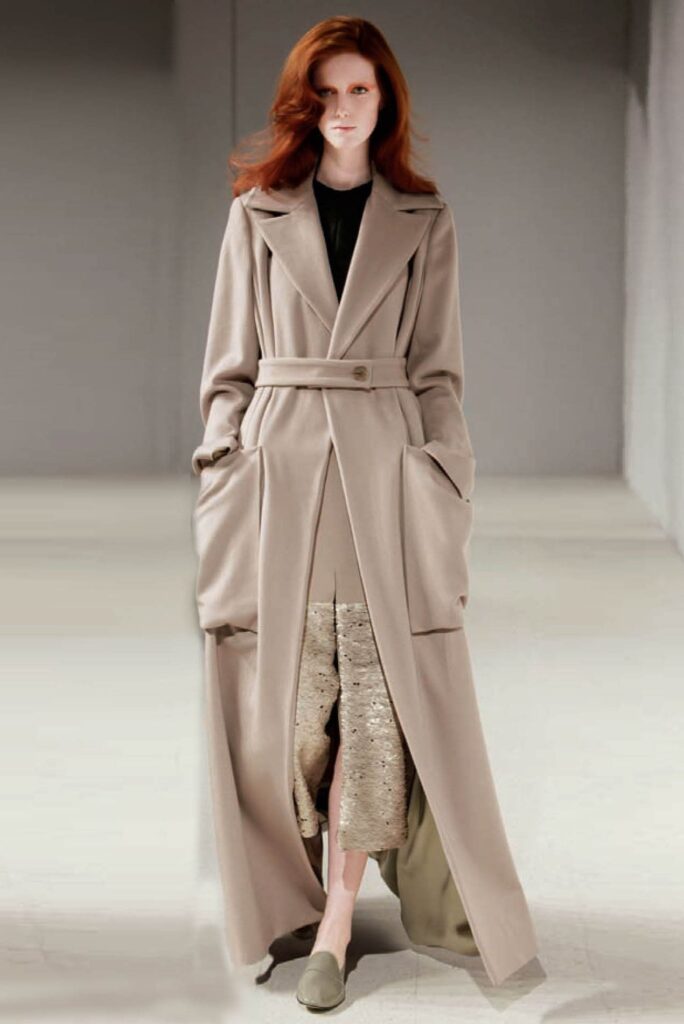 A model wearing a long beige belted coat over a textured skirt and matching shoes on a runway.