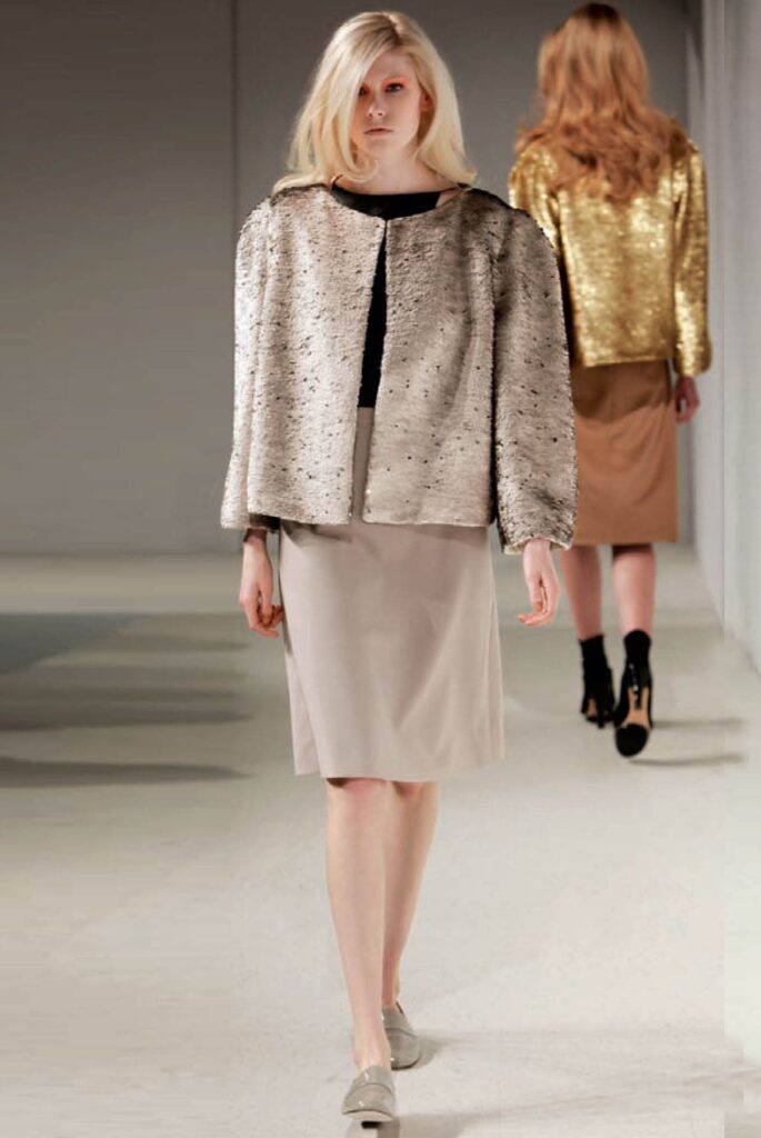 Model in a textured beige jacket and skirt walks a runway; another model in gold is behind her.