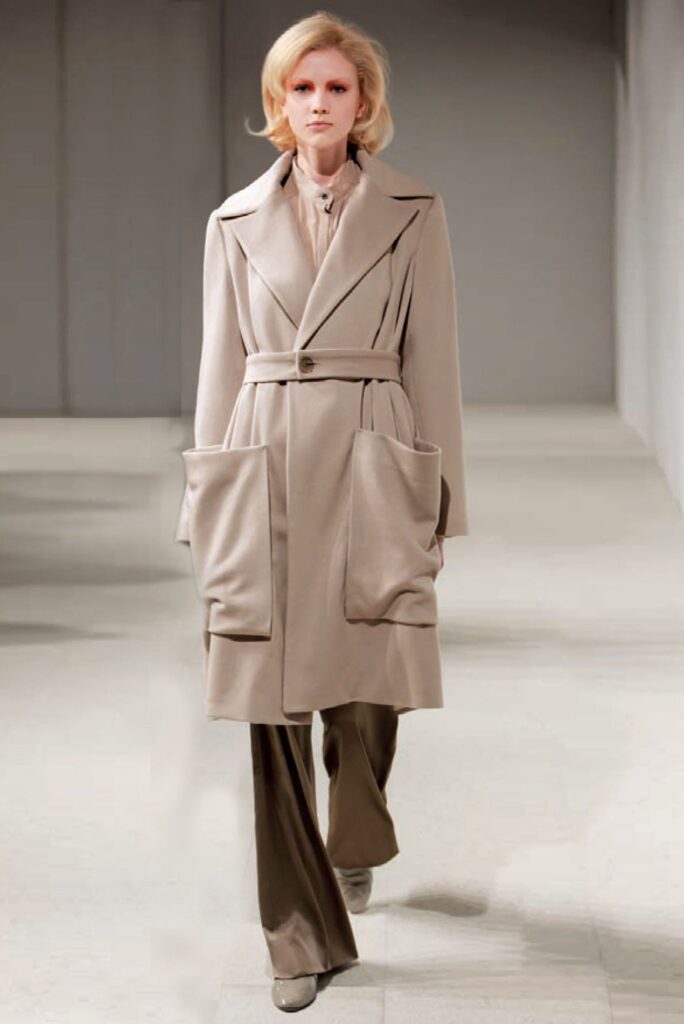 Person wearing a long beige belted coat with large pockets, wide-leg pants, and heeled shoes on a runway.