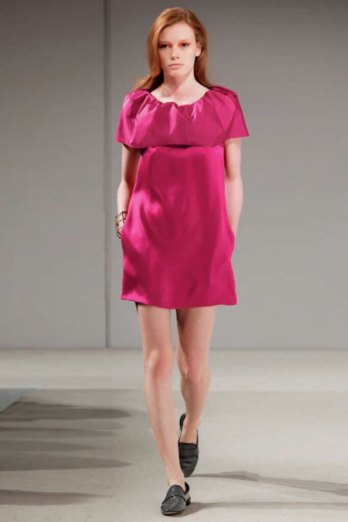 Model walks runway in a short, bright pink dress with cap sleeves and black loafers.