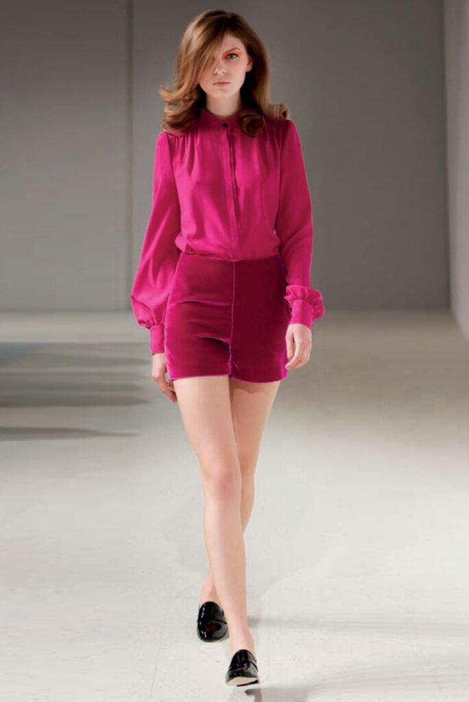 Woman walks runway in bright pink long-sleeve blouse and matching high-waisted shorts, with black loafers.