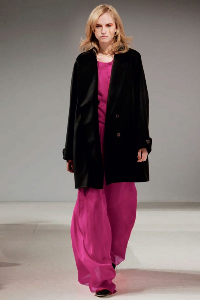 A woman walks the runway in a black coat over a bright pink blouse and wide-legged pink pants.