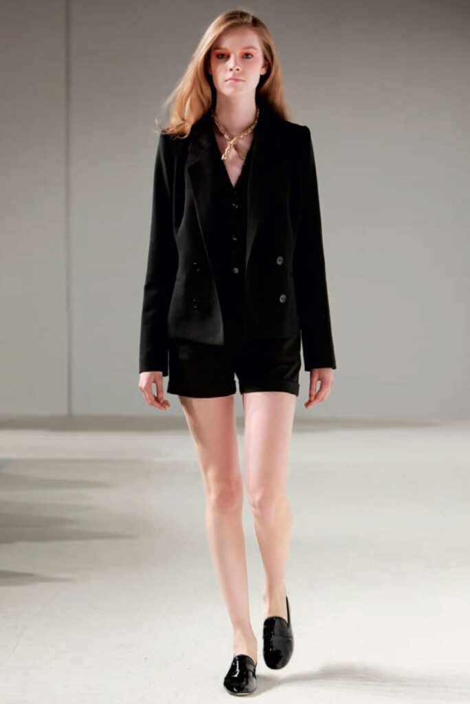 A person models a black blazer, black shorts, and black loafers on a runway with a neutral background.