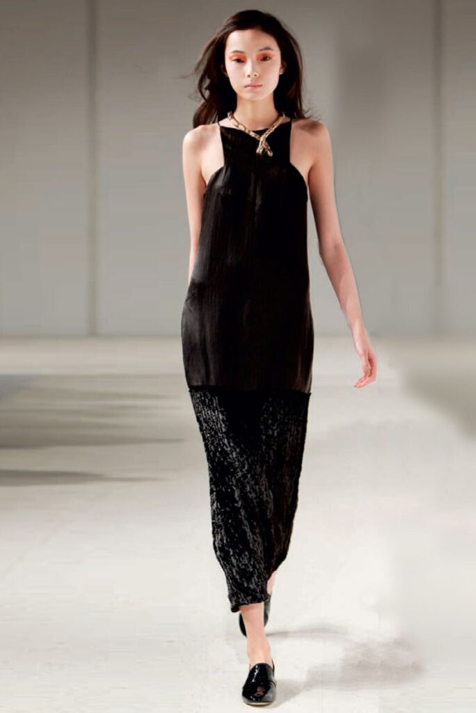 Woman on runway wears sleeveless black tunic dress over textured black pants, with black shoes.