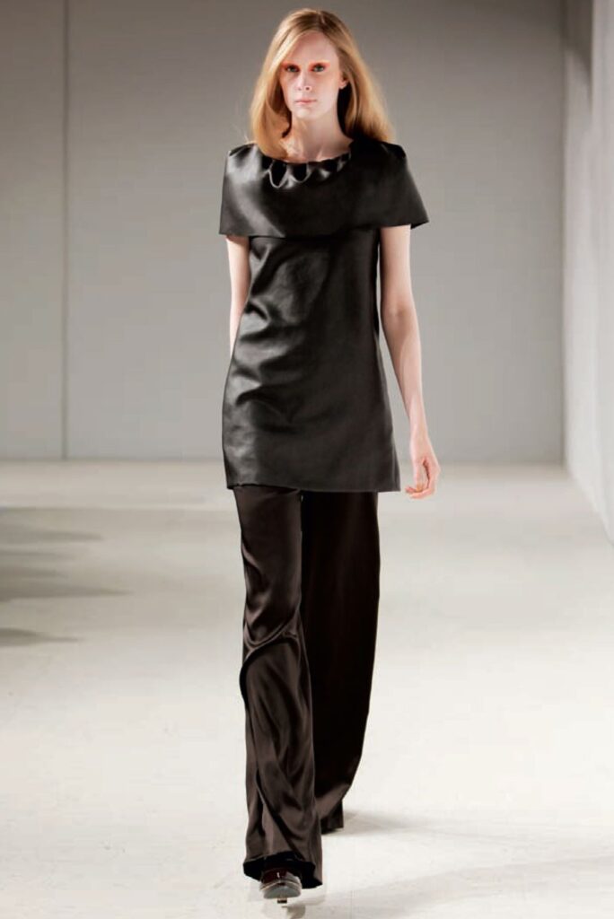 A model walks a runway in a short black dress layered over loose black pants and open-toe shoes.