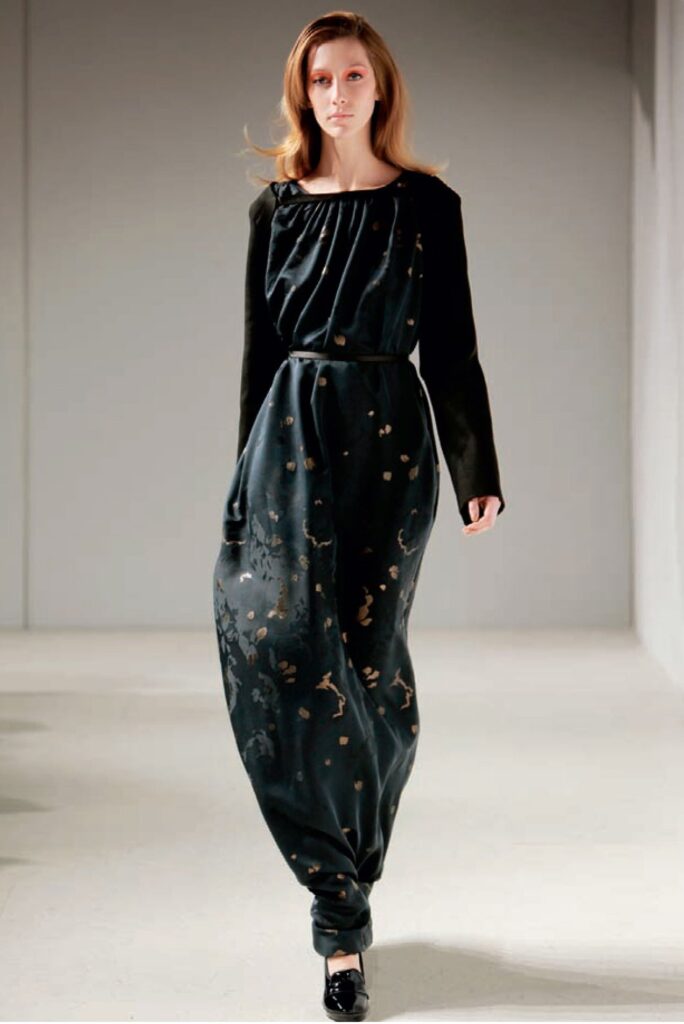 Model in a long, dark, off-shoulder dress with gold accents, walking on a plain, light runway.