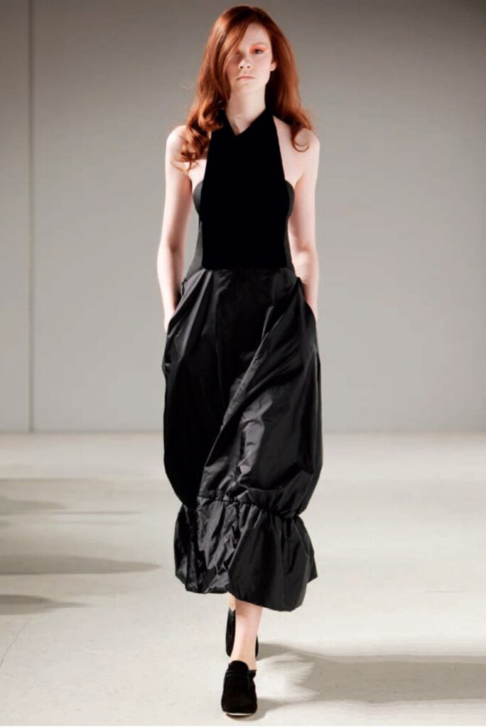 Woman with long red hair wears a sleeveless black dress and black shoes, walking on a light-colored floor.