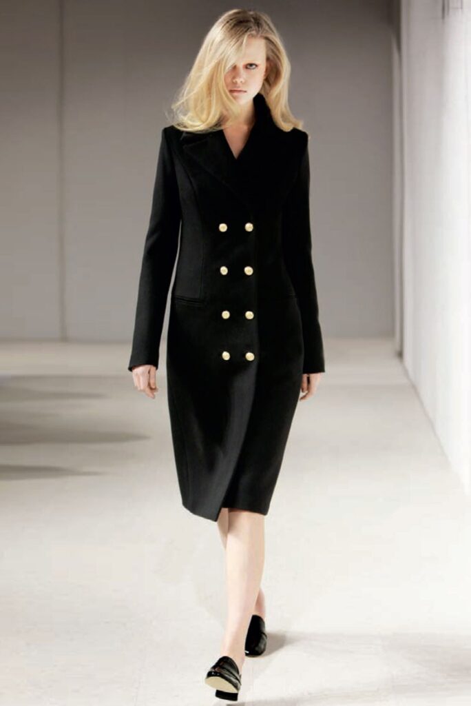 Woman wearing a black, double-breasted coat dress with gold buttons walks on a runway.