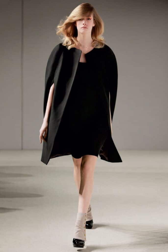 Woman walking runway in a black cape dress, beige socks, and black heels, with light wavy hair.