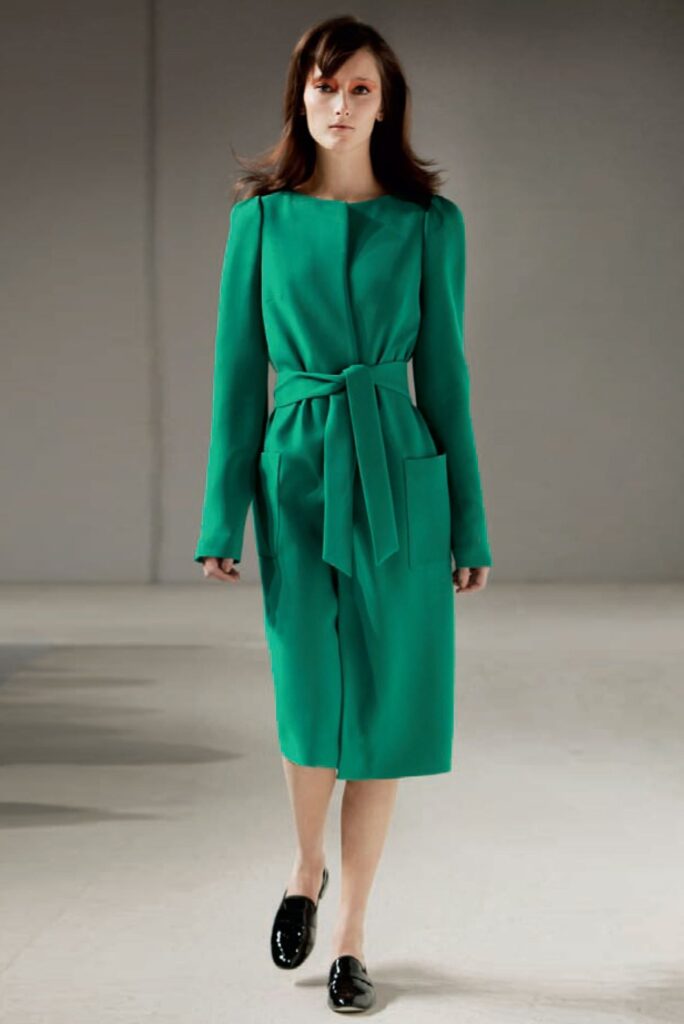 Woman in a bright green belted dress and black shoes walking on a runway with a neutral background.