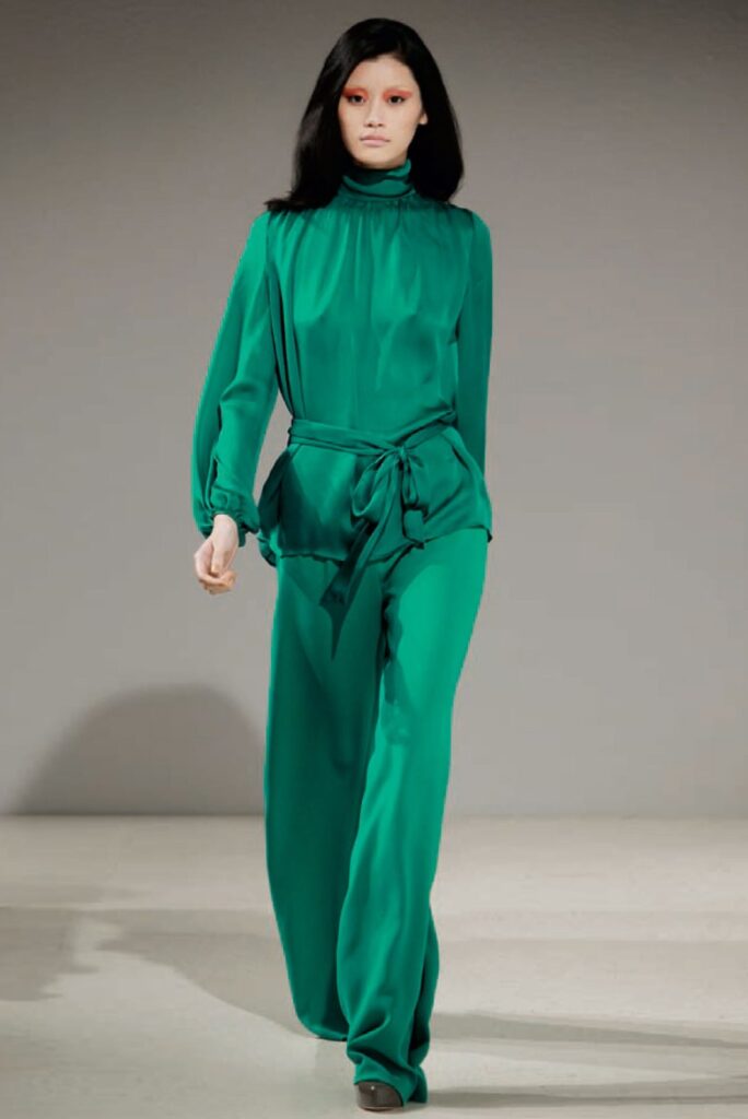 Woman in a flowing emerald green long-sleeve jumpsuit walks on a runway against a plain background.