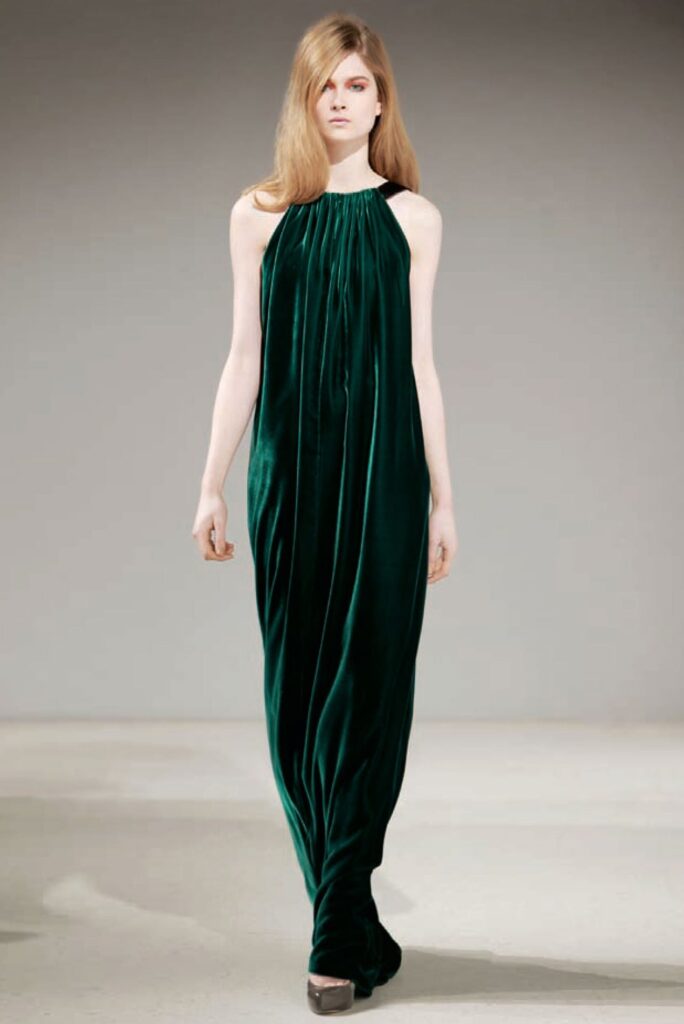 Model in a dark green, floor-length, sleeveless velvet gown with a gathered neckline walks a runway.