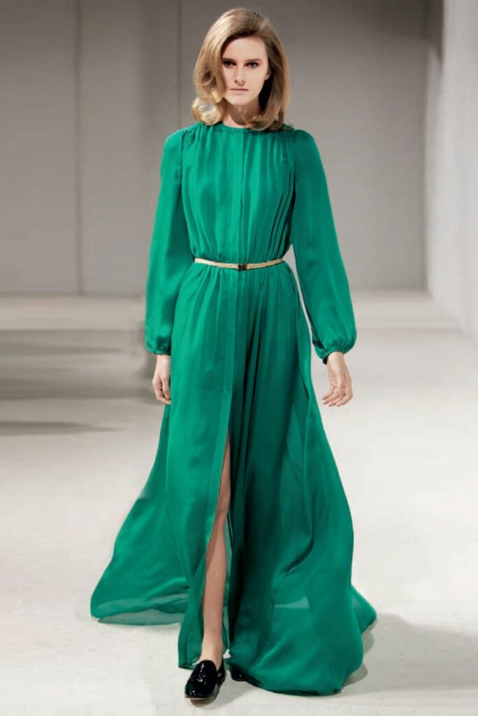 Woman in a long green dress with a gold belt, walking indoors on a light-colored floor.