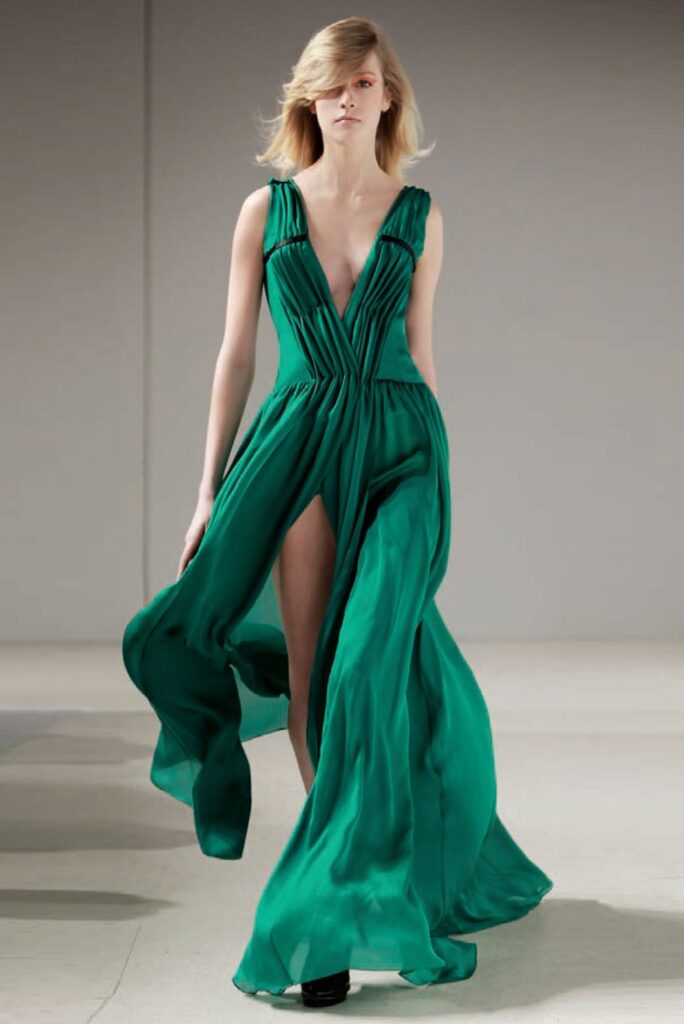 Model in a flowing, deep green gown with a plunging neckline and high slit, walking on a runway.