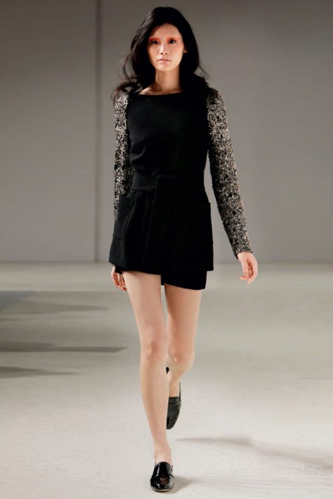 Woman modeling a black dress with patterned long sleeves and black shoes on a runway.