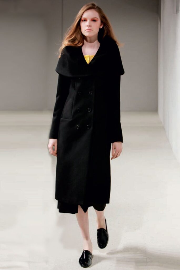 A person models a long black coat and black shoes, walking on a minimalist indoor runway.