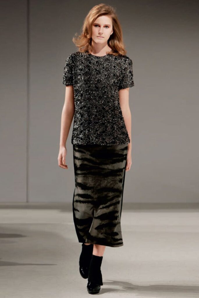 A model walks in a short-sleeve textured top and a long, patterned skirt with black ankle boots.