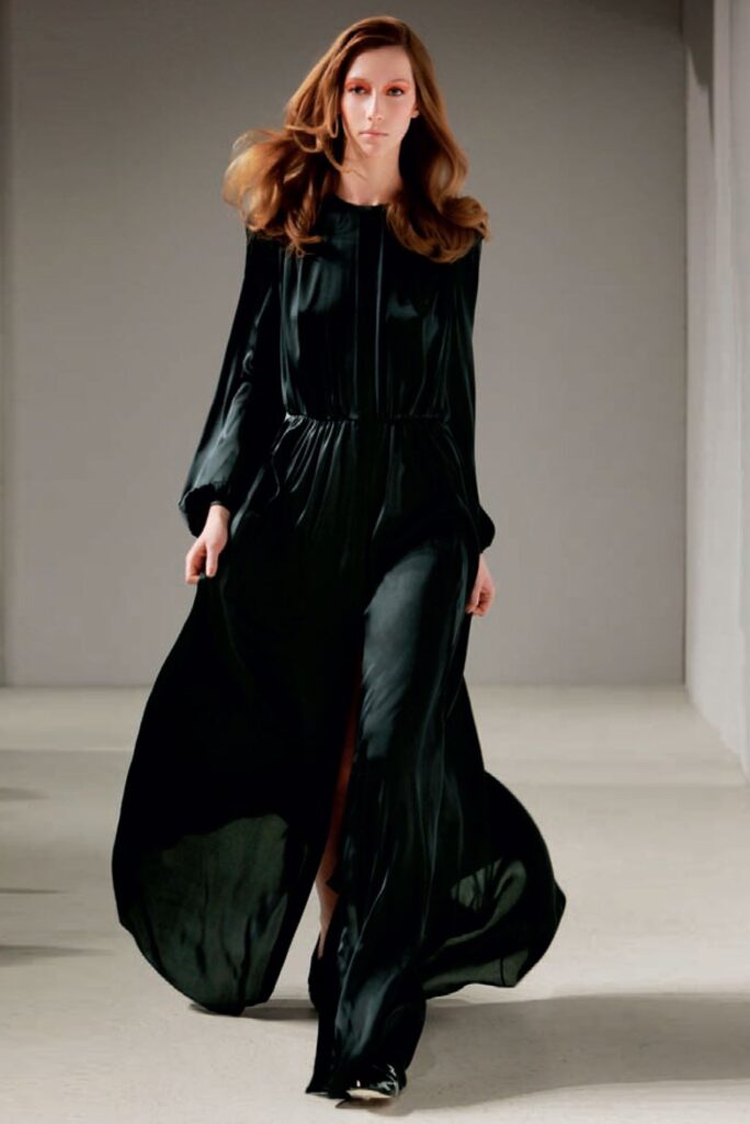Woman walking in a flowing, long-sleeved black dress on a runway in a minimalist setting.
