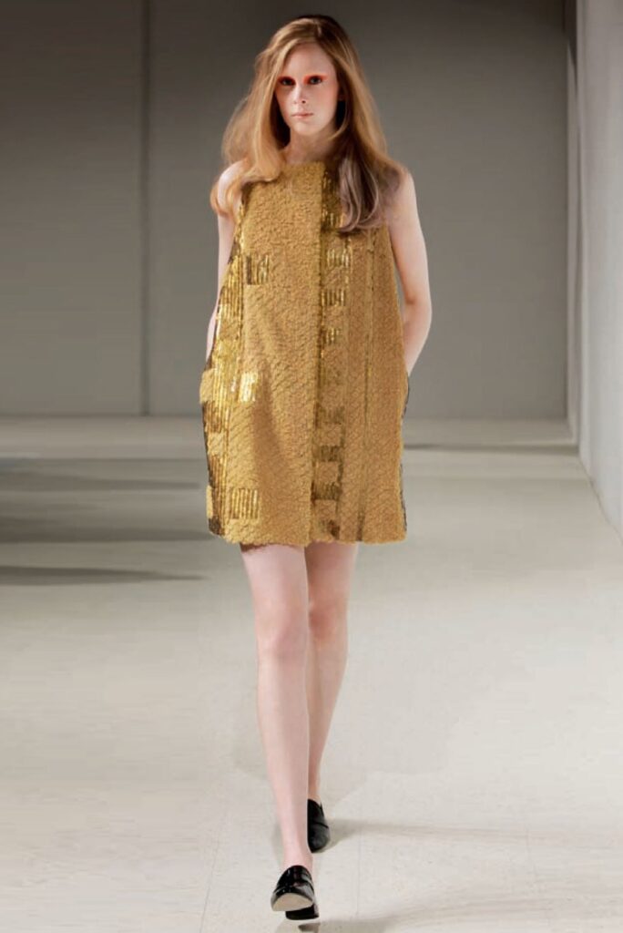 Model walks on runway wearing a gold textured sleeveless dress and black shoes, hands in dress pockets.