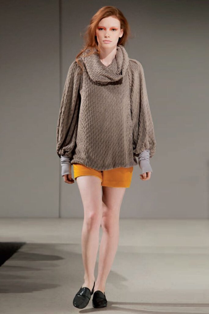 Model in a loose brown sweater with balloon sleeves, yellow shorts, and black shoes walking on a runway.