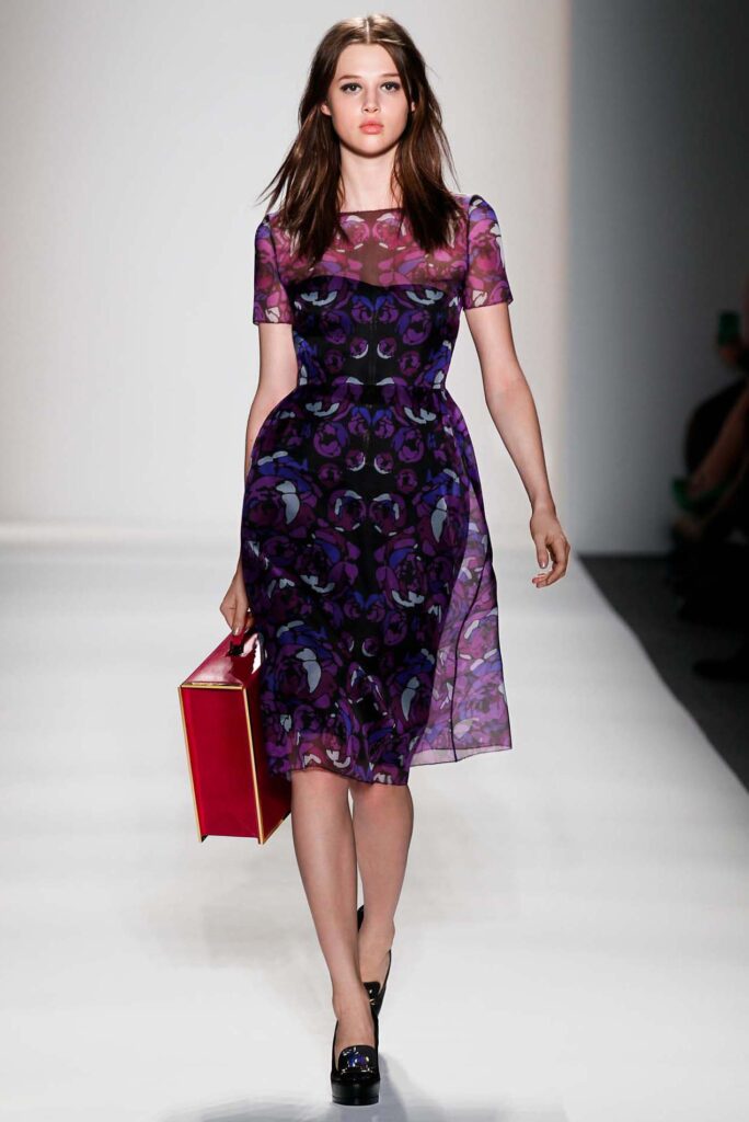 Model in a purple floral dress walking the runway, carrying a red box-shaped handbag.
