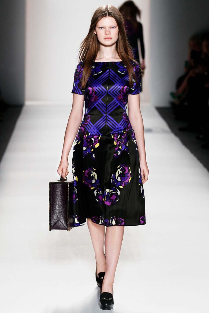 Model walking runway in a geometric purple and black dress, holding a rectangular black handbag.