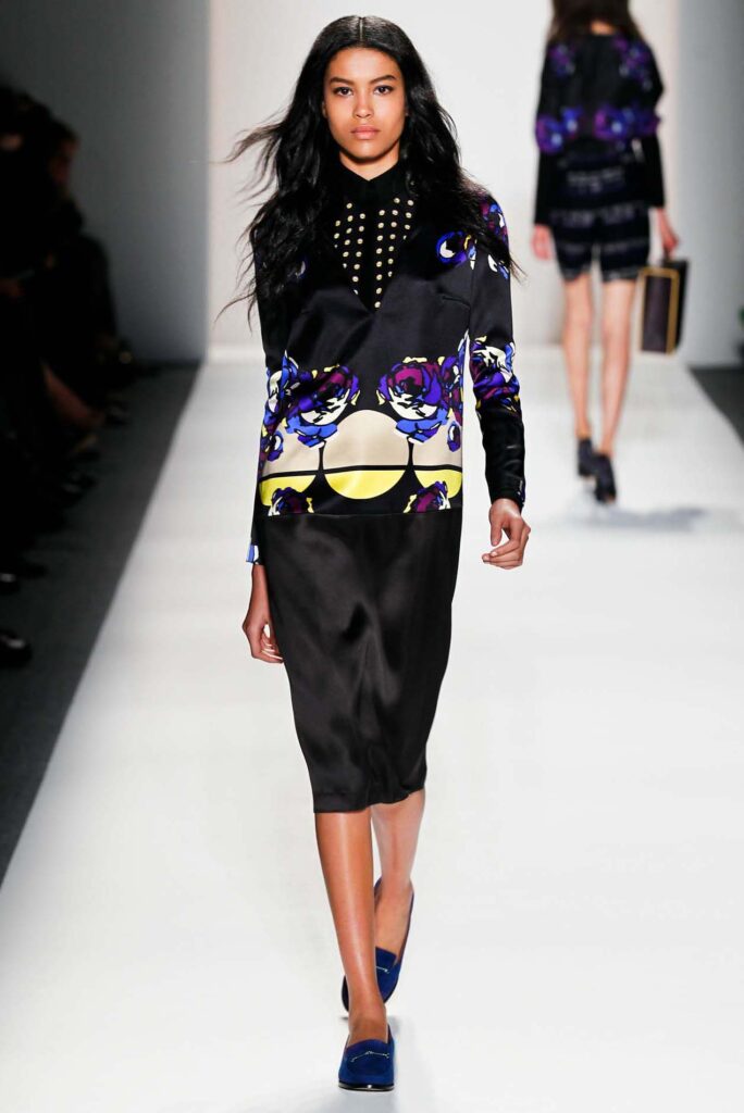 Model walks runway in a black dress with bold purple, yellow, and white floral patterns and blue shoes.