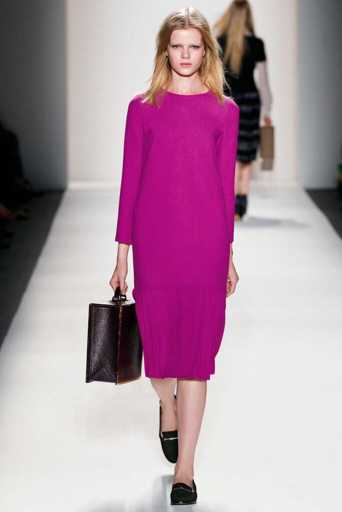 Model in a long-sleeve fuchsia dress walks runway holding a boxy handbag, wearing black loafers.