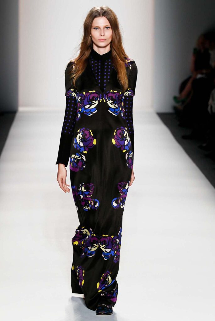 Model walks runway in a long black dress with bold purple, blue, and yellow floral prints, long sleeves.