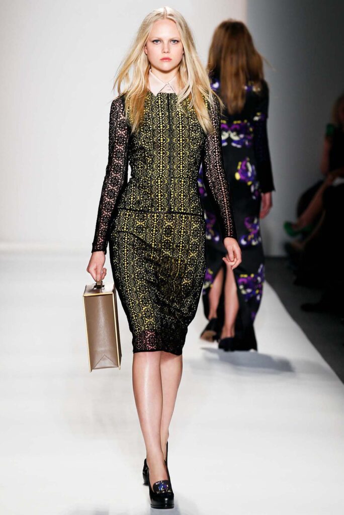 Model in a black and gold lace dress carrying a boxy bag walks down a fashion runway.