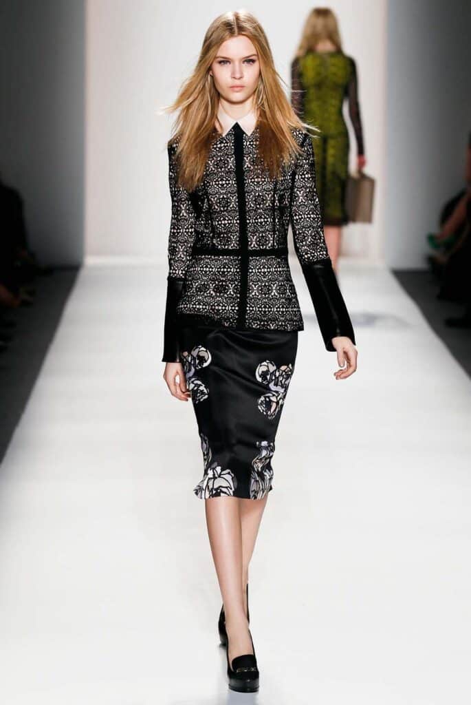 A model walks a runway in a patterned black jacket and skirt with floral designs, wearing black heels.