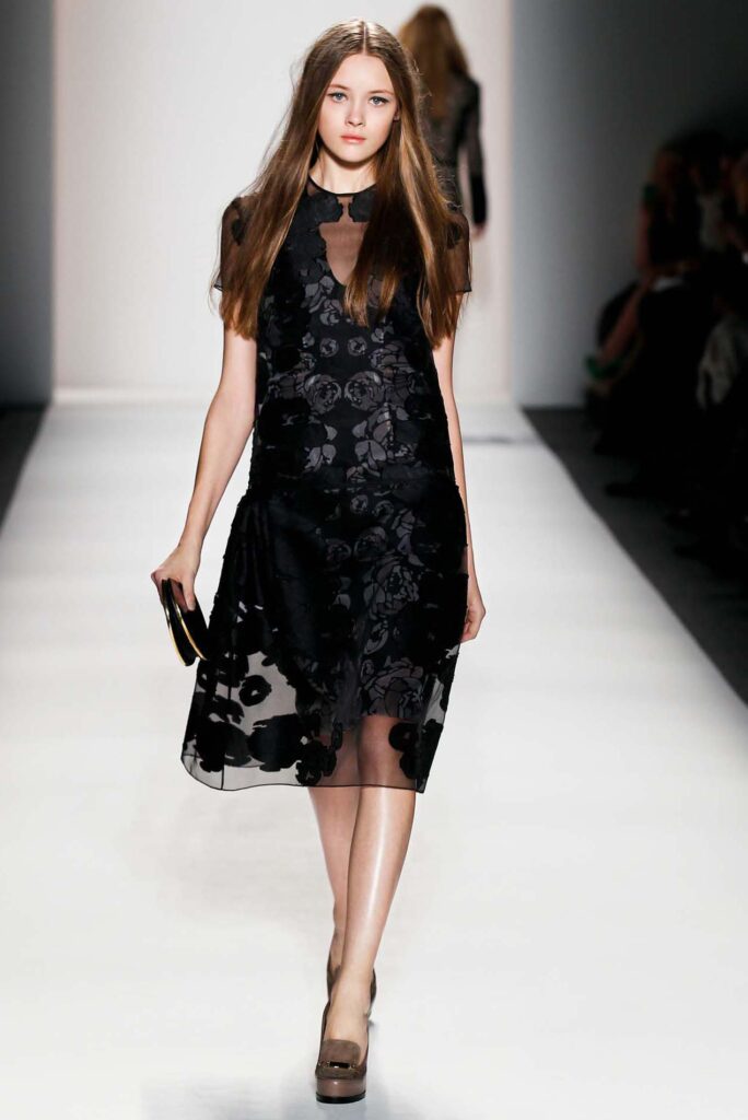 Model walks runway in a black floral dress with sheer overlay, holding a clutch, on a white fashion show stage.