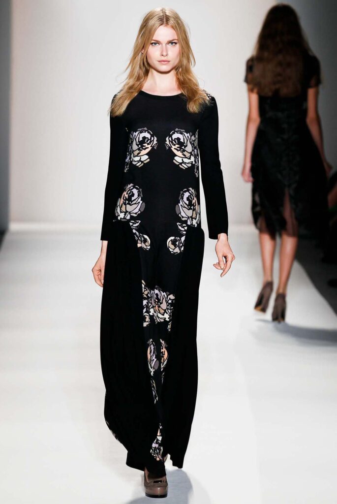 Model in a long black floral dress walks down a runway; another model is visible in the background.