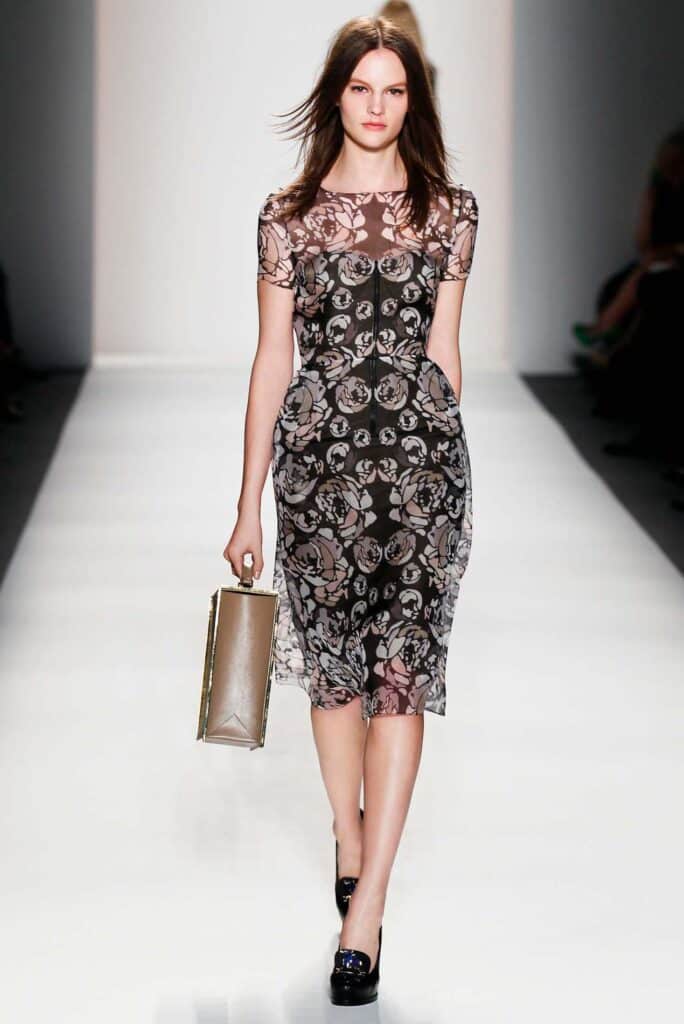 Model in a sheer floral dress walks runway, holding a clutch, with a white background.