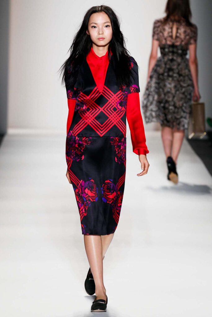 Model walking runway in a red and black geometric dress with floral patterns, black flats, and loose hair.