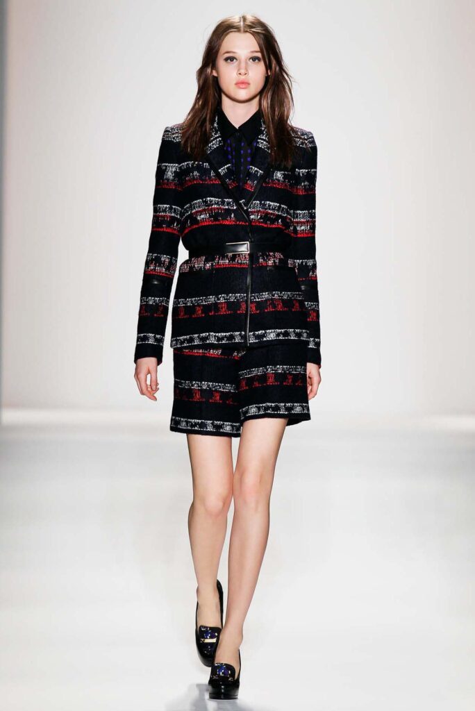 Woman walks runway in a patterned black blazer and matching skirt, wearing black shoes, on a white background.
